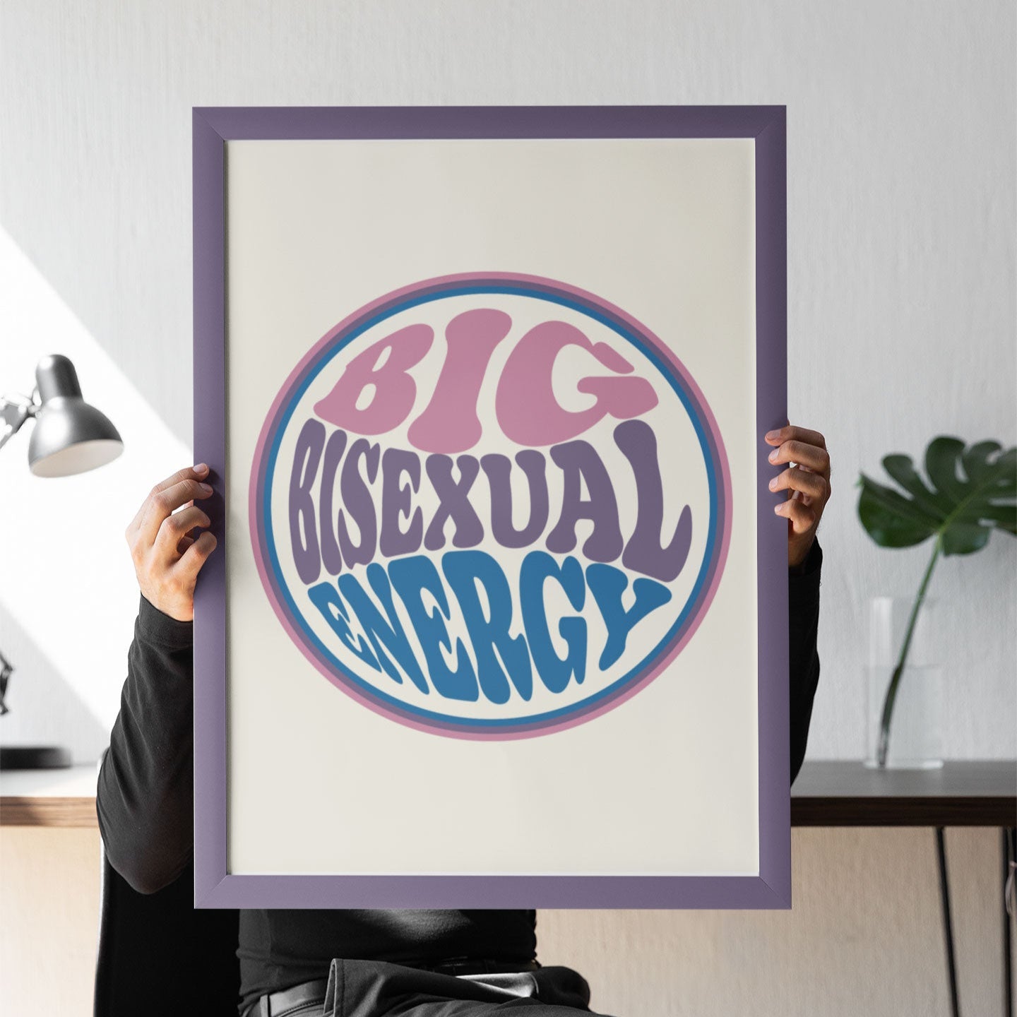 A person in black clothing sits on a modern chair, holding a large framed Big Bisexual Energy print in front of their face. The background includes a minimalist desk with a lamp and a plant.