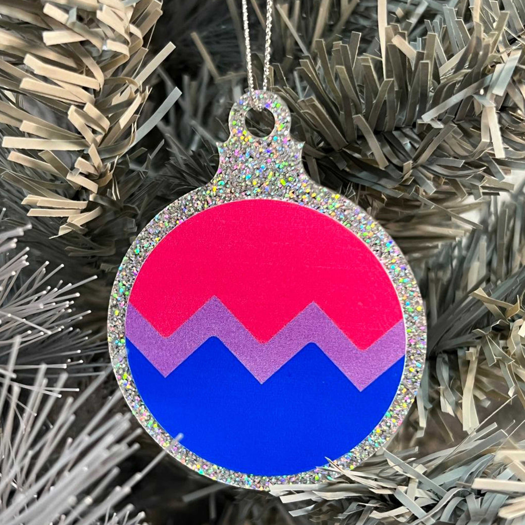 Close up of a white Christmas tree displaying a silver glitter bauble featuring a classic zig zag design in the colours of the bisexual pride flag.