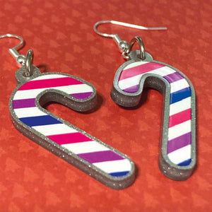 Silver glitter candy cane dangle earrings lay on a red christmas tree paper background. The stripes on the candy cane are those of the Bisexual flag; pink, purple, and blue.