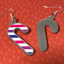 Load image into Gallery viewer, Silver glitter candy cane dangle earrings lay on a red christmas tree paper background. The stripes on the candy cane are those of the Bisexual flag; pink, purple, and blue. One of the earrings is turned over to show the reverse, which is plain silver glitter.
