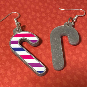 Silver glitter candy cane dangle earrings lay on a red christmas tree paper background. The stripes on the candy cane are those of the Bisexual flag; pink, purple, and blue. One of the earrings is turned over to show the reverse, which is plain silver glitter.