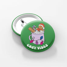 Load image into Gallery viewer, Close up of a green 38mm badge featuring retro text reading &#39;cosy vibes&#39;. The image shows a mug of hot chocolate with marshmallows and a gingerbread man wearing a Santa hat. A candy cane in the mug and lights around the mug are the colours of the bisexual flag.
