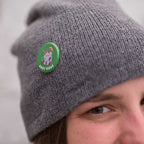 Young woman wearing a grey knitted hat with a badge pinned to it. The badge is a green 38mm badge featuring retro text reading 'cosy vibes'. The badge shows a mug of hot chocolate with marshmallows and a gingerbread man wearing a Santa hat. A candy cane in the mug and lights around the mug are the colours of the bisexual flag.