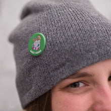 Load image into Gallery viewer, Young woman wearing a grey knitted hat with a badge pinned to it. The badge is a green 38mm badge featuring retro text reading &#39;cosy vibes&#39;. The badge shows a mug of hot chocolate with marshmallows and a gingerbread man wearing a Santa hat. A candy cane in the mug and lights around the mug are the colours of the bisexual flag.