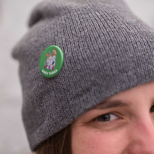 Young woman wearing a grey knitted hat with a badge pinned to it. The badge is a green 38mm badge featuring retro text reading 'cosy vibes'. The badge shows a mug of hot chocolate with marshmallows and a gingerbread man wearing a Santa hat. A candy cane in the mug and lights around the mug are the colours of the bisexual flag.