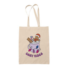 Load image into Gallery viewer, Natural tote bag featuring retro text reading &#39;cosy vibes&#39;. The image shows a mug of hot chocolate with marshmallows and a gingerbread man wearing a Santa hat. A candy cane in the mug and lights around the mug are the colours of the bisexual pride flag.