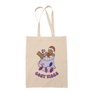 Natural tote bag featuring retro text reading 'cosy vibes'. The image shows a mug of hot chocolate with marshmallows and a gingerbread man wearing a Santa hat. A candy cane in the mug and lights around the mug are the colours of the bisexual pride flag.