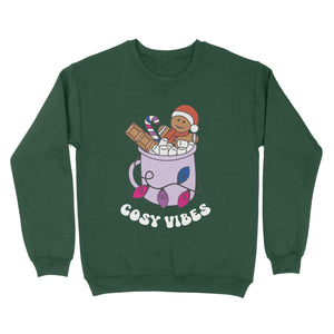 Bottle Green sweatshirt featuring retro text reading 'cosy vibes'. The image shows a mug of hot chocolate with marshmallows and a gingerbread man wearing a Santa hat. A candy cane in the mug and lights around the mug are the colours of the bisexual pride flag.