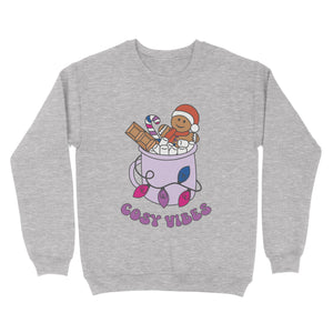 Heather Grey sweatshirt featuring retro text reading 'cosy vibes'. The image shows a mug of hot chocolate with marshmallows and a gingerbread man wearing a Santa hat. A candy cane in the mug and lights around the mug are the colours of the bisexual pride flag.
