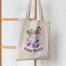 Load image into Gallery viewer, Natural tote bag featuring retro text reading &#39;cosy vibes&#39;. The image shows a mug of hot chocolate with marshmallows and a gingerbread man wearing a Santa hat. A candy cane in the mug and lights around the mug are the colours of the bisexual pride flag hanging from some wooden ladders.