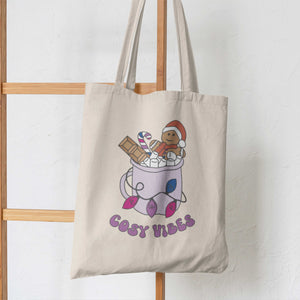 Natural tote bag featuring retro text reading 'cosy vibes'. The image shows a mug of hot chocolate with marshmallows and a gingerbread man wearing a Santa hat. A candy cane in the mug and lights around the mug are the colours of the bisexual pride flag hanging from some wooden ladders.