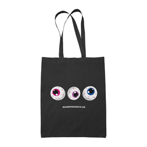 A black tote bag featuring three cartoonish eyeballs in a row. The eyeballs display different colours: one with a pink iris, one with a purple iris, and one with a blue iris. The text "RAINBOWANDCO.UK" is positioned below the eyeballs.