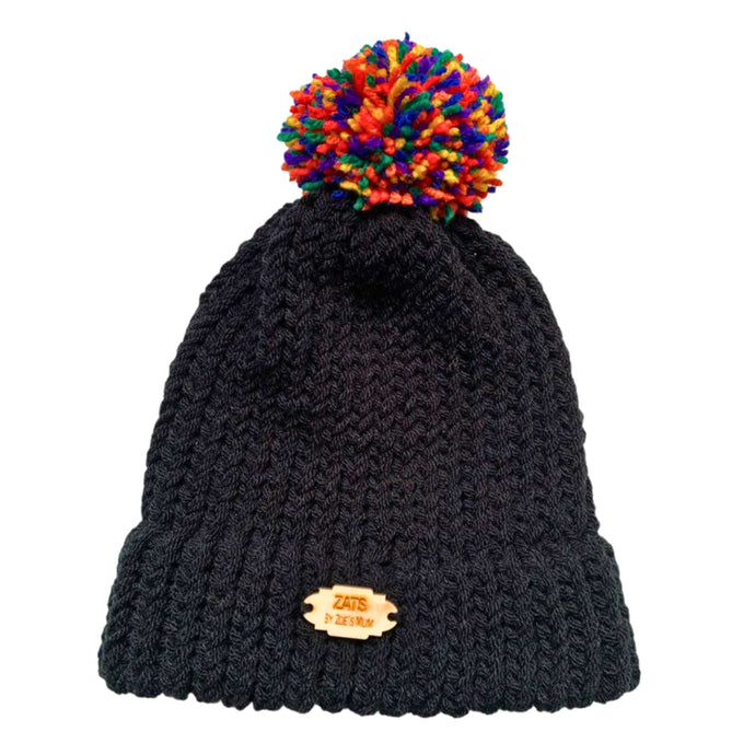 Black knitted bobble hat with a pom pom in the colours of the rainbow pride flag laying flat.