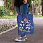 Person carrying a royal blue cotton tote bag printed with the slogan "Buy Like You Give A Damn" and "Your Money Is Your Vote" while walking along a tree-lined path, showcasing the bag in everyday use.