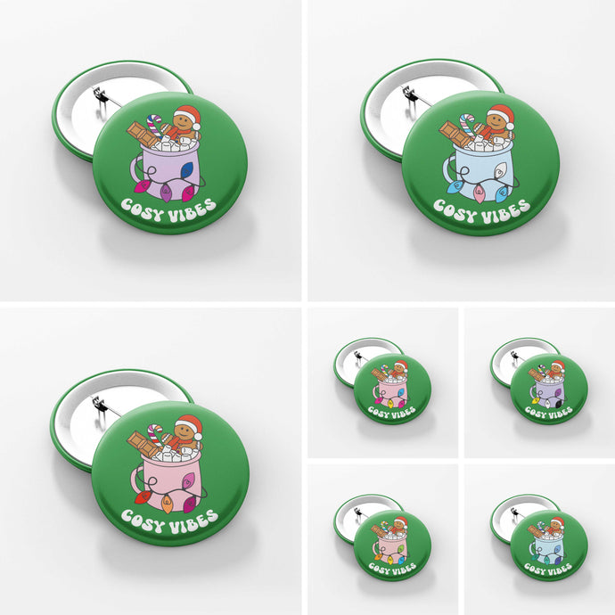 Image is split into seven squares, each one showing a close up of a green 38mm badge featuring retro text reading 'cosy vibes'. Each badge shows a mug of hot chocolate with marshmallows and a gingerbread man wearing a Santa hat. A candy cane in the mug and lights around the mug are the colours of different pride flags.
