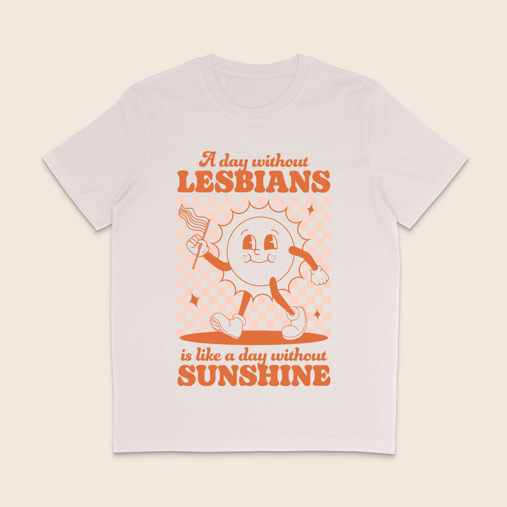 A vintage white short-sleeve crewneck t-shirt with a retro-style orange graphic on the front. The graphic features a cartoon sun character holding a flag and the text 'A day without LESBIANS is like a day without SUNSHINE'.