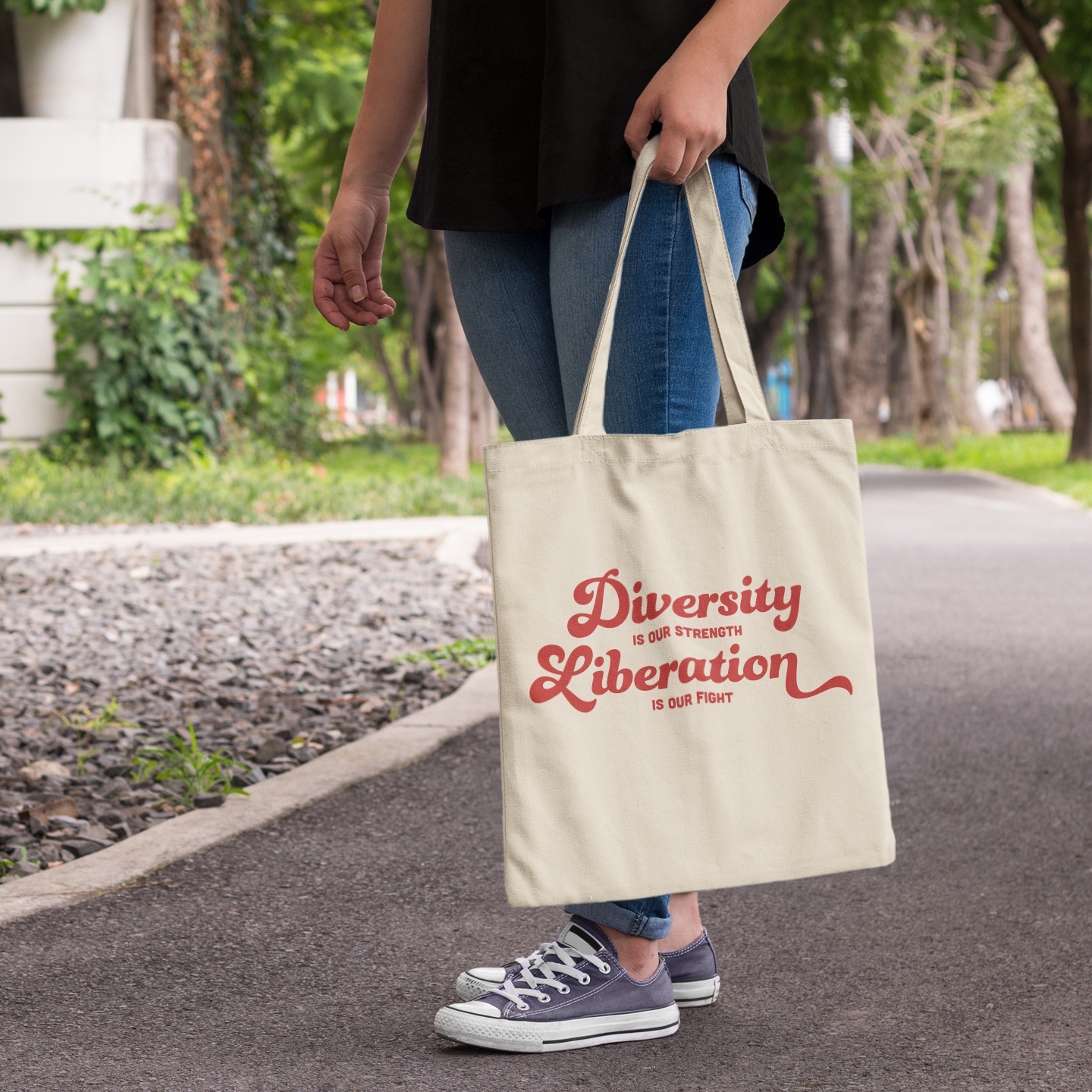 A natural canvas tote bag carried by someone walking outdoors on a park path. The bag displays the bold red slogan “Diversity is our Strength, Liberation is our Fight” in a retro-style script font.
