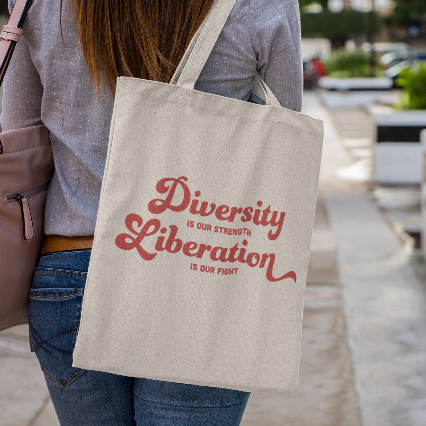 A natural canvas tote bag worn over the shoulder of a woman in a grey spotted shirt. The design reads “Diversity is our Strength, Liberation is our Fight” in bold red lettering across the front.