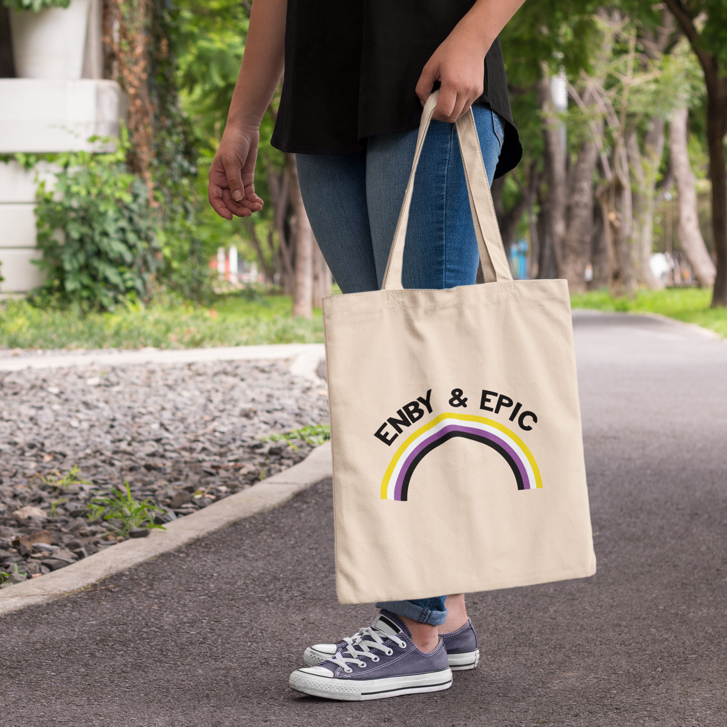 Natural cotton tote bag with the slogan “Enby & Epic” and a rainbow arc in non-binary pride colours, shown worn outdoors