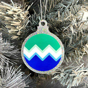 Close up of a white Christmas tree displaying a silver glitter bauble featuring a classic zig zag design in the colours of the gay pride flag.