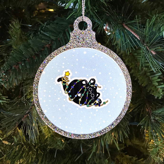 A glittery Christmas bauble with a black cat tangled in bright fairy lights in the colours of the gay pride flag. The cat, looking cute and festive, lies against a snowy background, hanging on a Christmas tree.