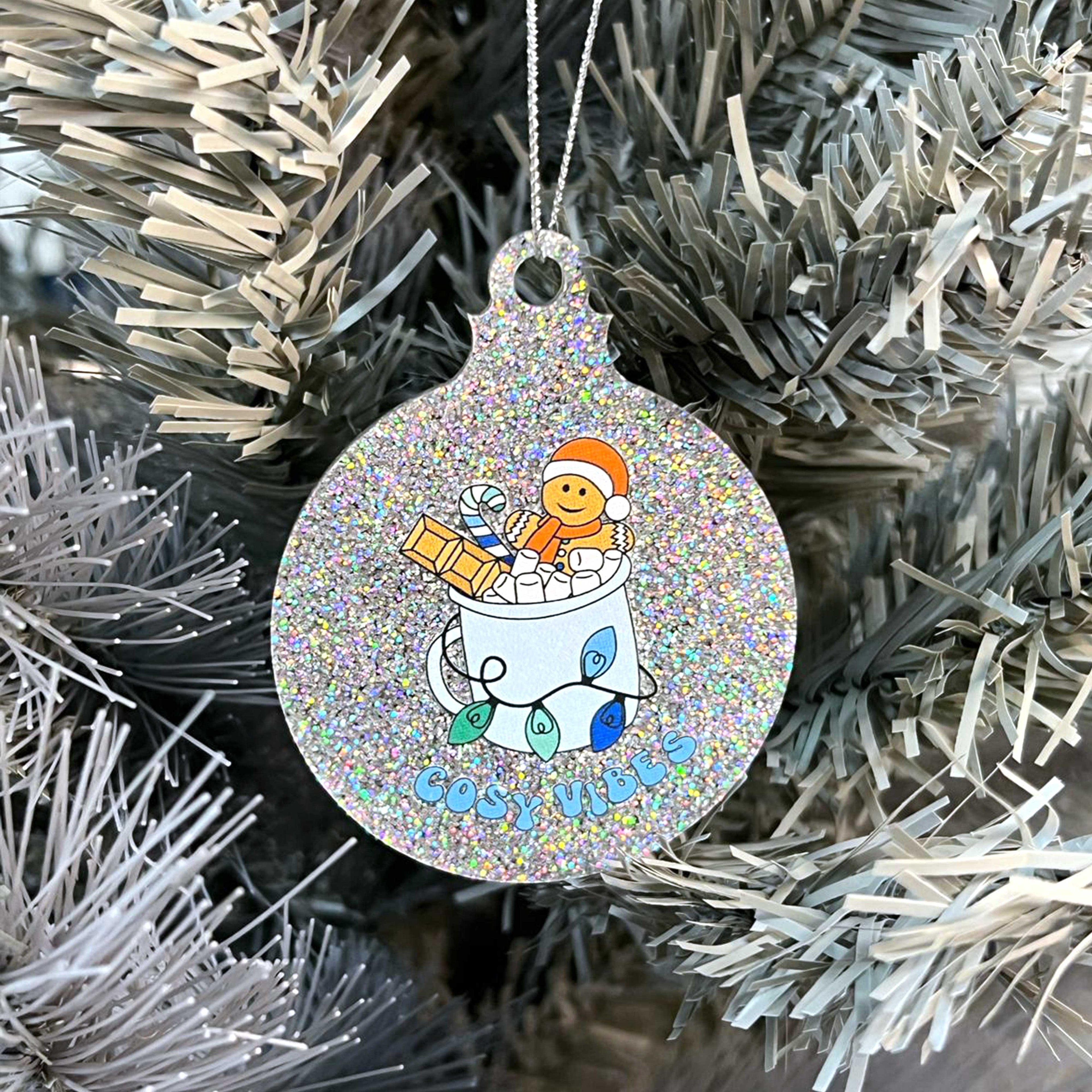 Close up of a white Christmas tree displaying a silver glitter bauble featuring retro text reading 'cosy vibes'. The image shows a mug of hot chocolate with marshmallows and a gingerbread man wearing a Santa hat. A candy cane in the mug and lights around the mug are the colours of the gay pride flag.