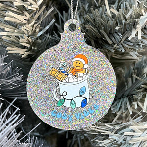 Close up of a white Christmas tree displaying a silver glitter bauble featuring retro text reading 'cosy vibes'. The image shows a mug of hot chocolate with marshmallows and a gingerbread man wearing a Santa hat. A candy cane in the mug and lights around the mug are the colours of the gay pride flag.