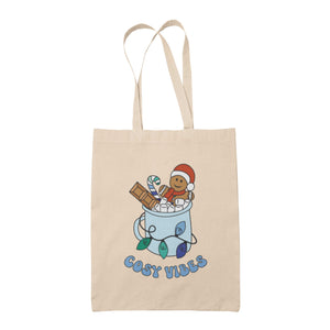 Natural tote bag featuring retro text reading 'cosy vibes'. The image shows a mug of hot chocolate with marshmallows and a gingerbread man wearing a Santa hat. A candy cane in the mug and lights around the mug are the colours of the gay pride flag.