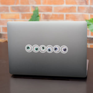 A row of six circular stickers with eyeball designs is aligned on the back of a grey laptop. The irises display a variety of colours, including different shades of green, blue, and purple representing the gay pride flag.