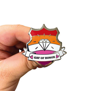 White hand holding up an enamel pin in the shape of a shield with a banner across it's front. Banner reads Gay of Honor. The background of the pin is the lesbian pride flag and there is a jewel icon in the centre. The jewel and banner are filled with white glitter.
