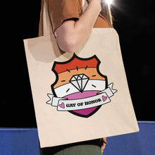 Load image into Gallery viewer, Close up of a white person with long hair carrying a natural coloured tote bag featuring a shield with a banner across it&#39;s front. Banner reads Gay of Honor. The background of the shield is the lesbian pride flag and there is a jewel icon in the centre.