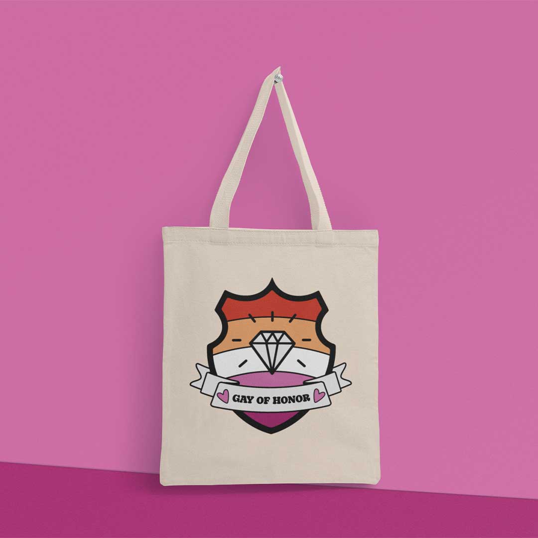 Natural coloured tote bag hanging from a peg on a pink wall. The bag features a shield with a banner across it's front. Banner reads Gay of Honor. The background of the shield is the lesbian pride flag and there is a jewel icon in the centre.