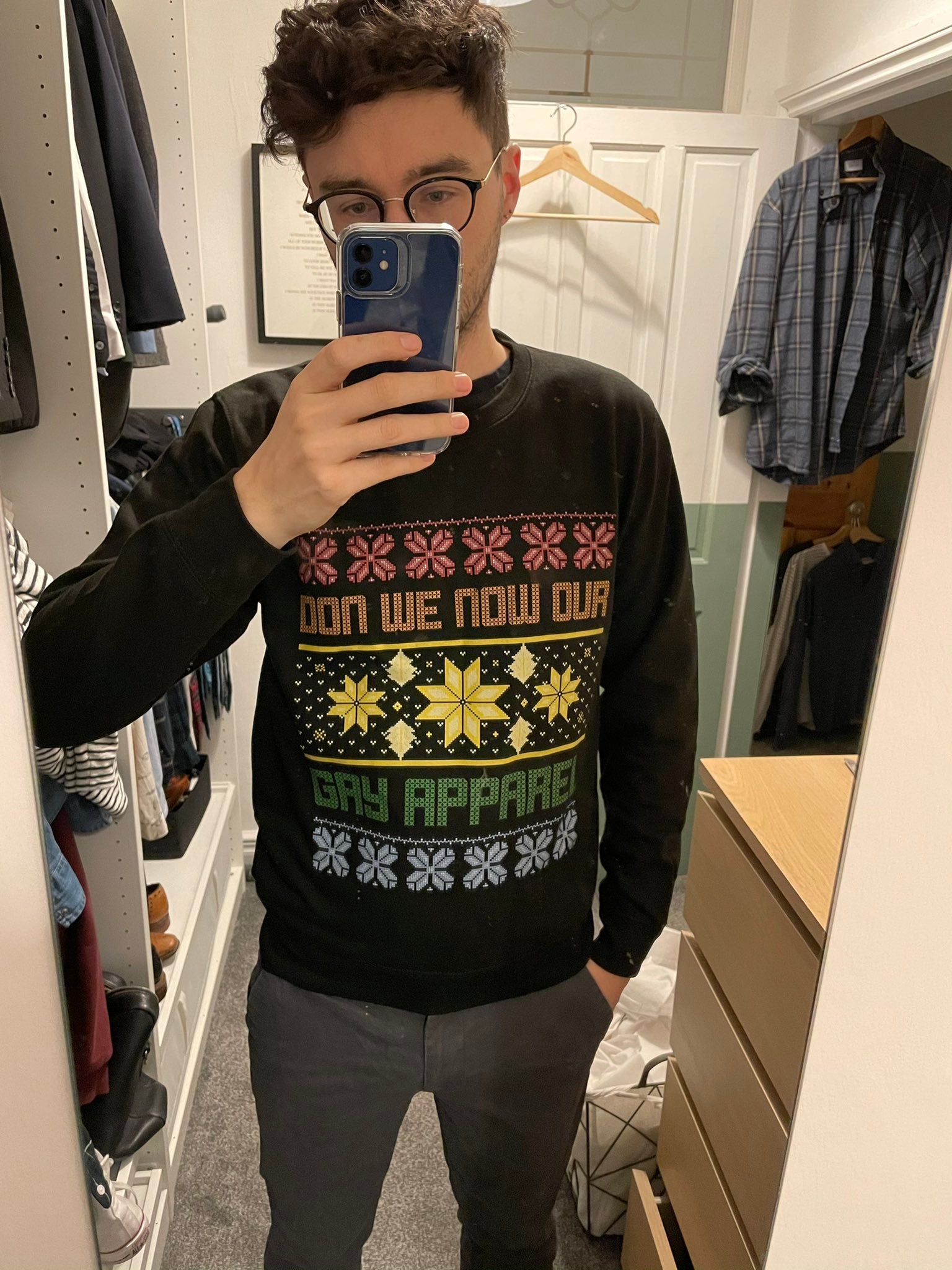 A man takes a mirror selfie in a closet wearing a black long-sleeve crewneck sweatshirt with a colorful Christmas-themed graphic that reads 'GAY APPAREL'.