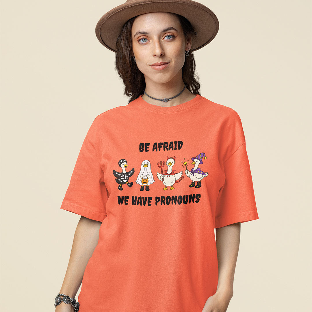 Model wearing a fiesta orange Be Afraid We Have Pronouns Halloween T-shirt featuring four geese dressed in spooky costumes.