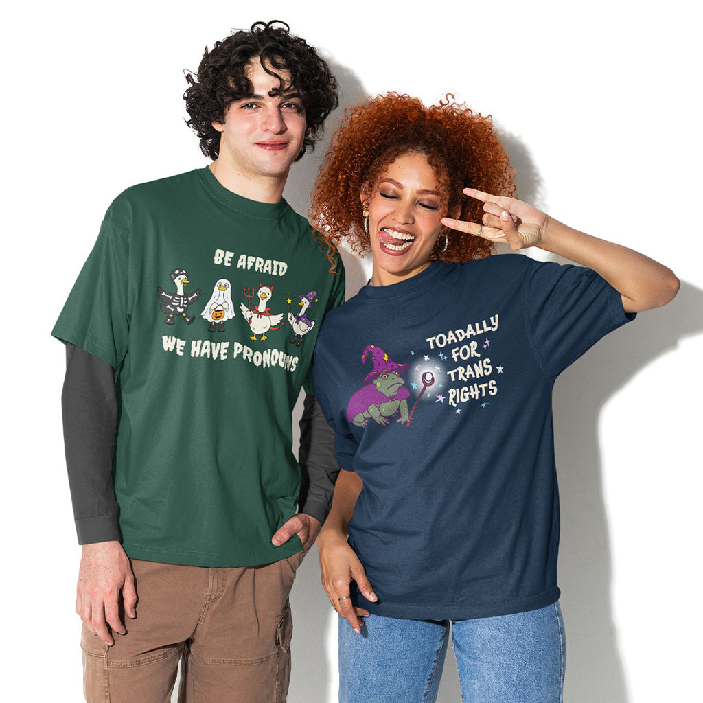 Two models wearing queer Halloween T-shirts featuring the Be Afraid We Have Pronouns geese design and Toadally for Trans Rights wizard toad design.