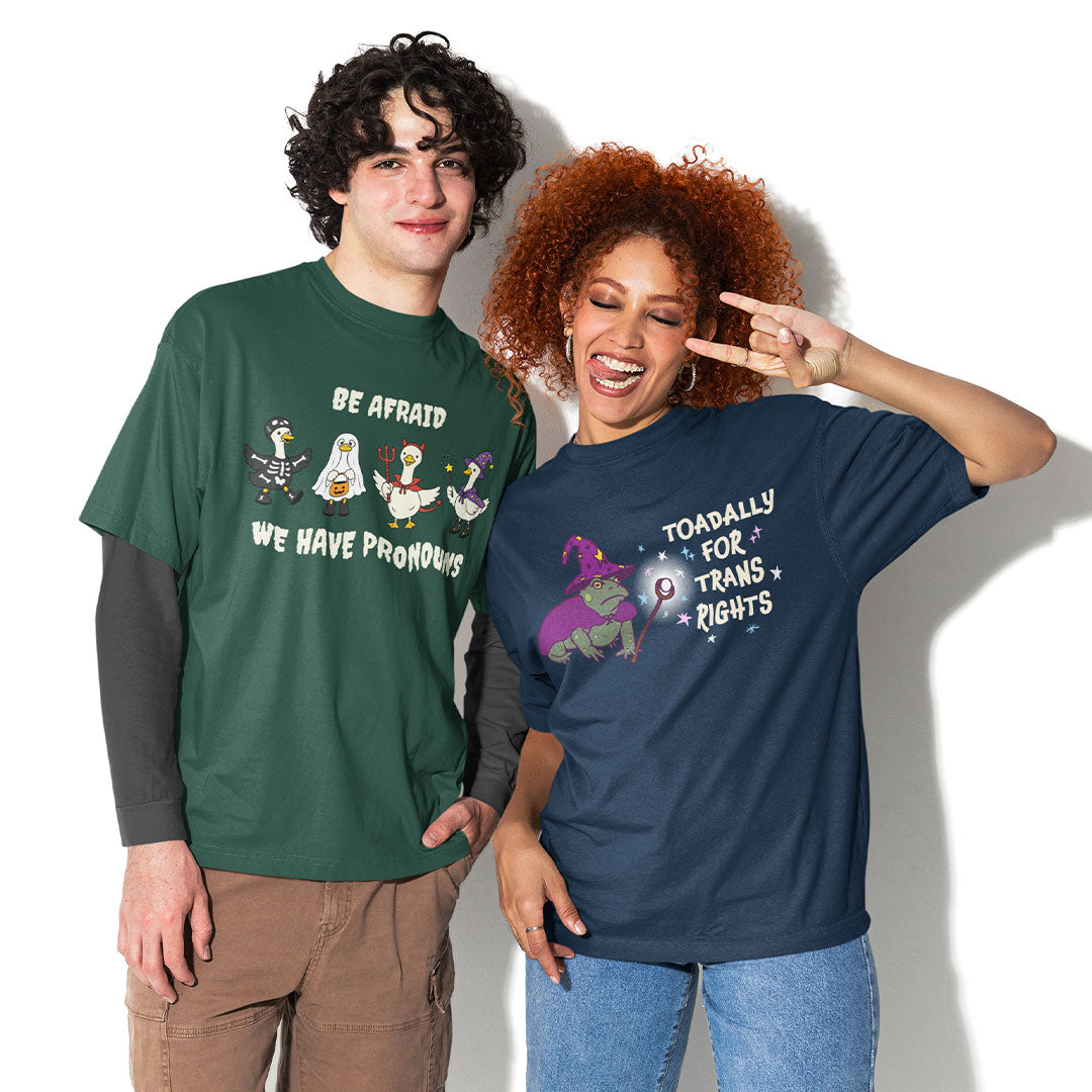 Two models wearing queer Halloween T-shirts featuring the Be Afraid We Have Pronouns geese design and Toadally for Trans Rights wizard toad design.