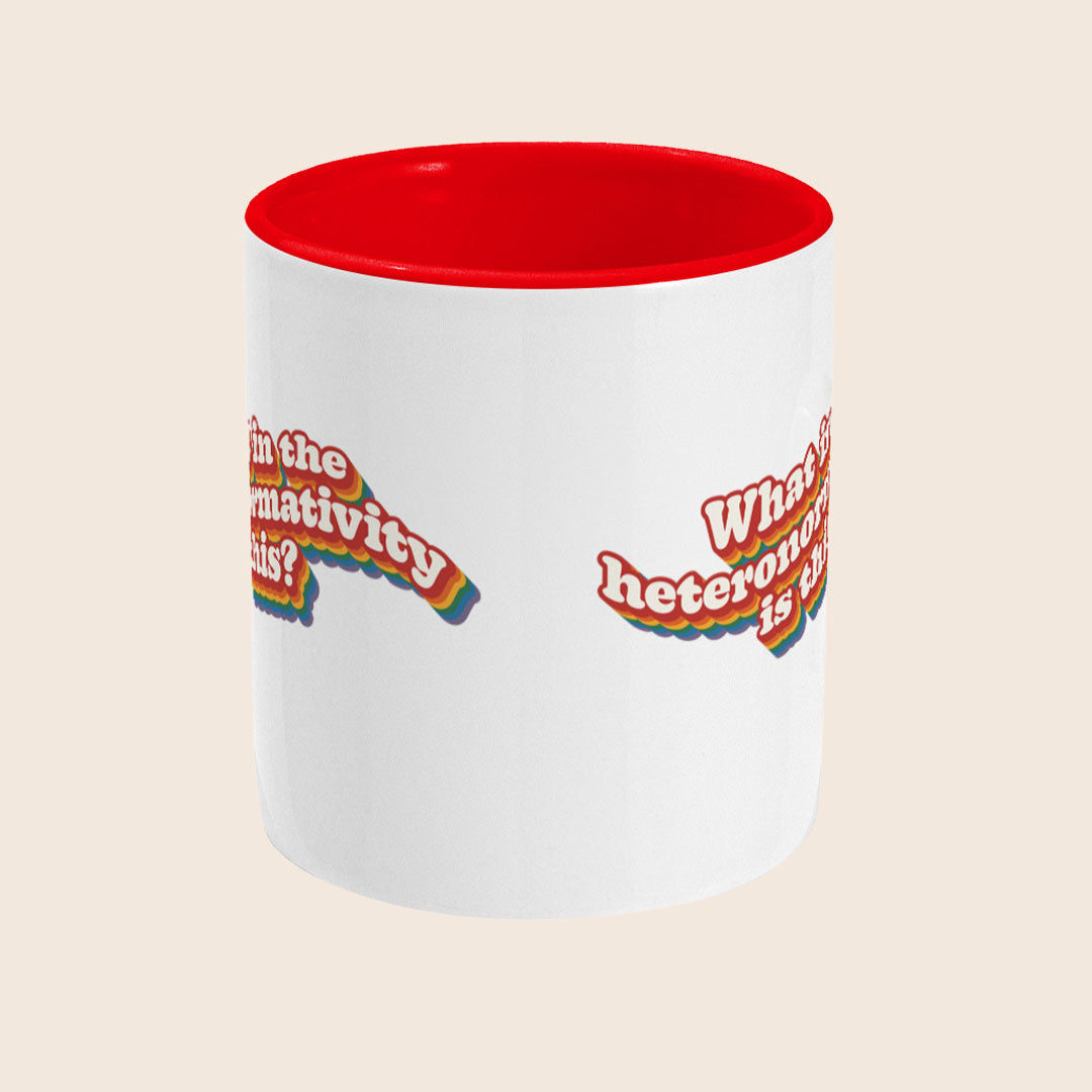 White ceramic mug with a red handle and red interior, featuring the phrase “What in the heteronormativity is this?” in rainbow-layered retro text across the front.