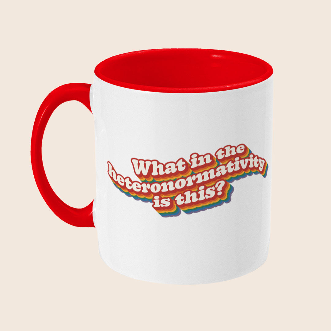 White ceramic mug with a red handle and red interior shown from a left angle, featuring the rainbow-layered slogan “What in the heteronormativity is this?”.