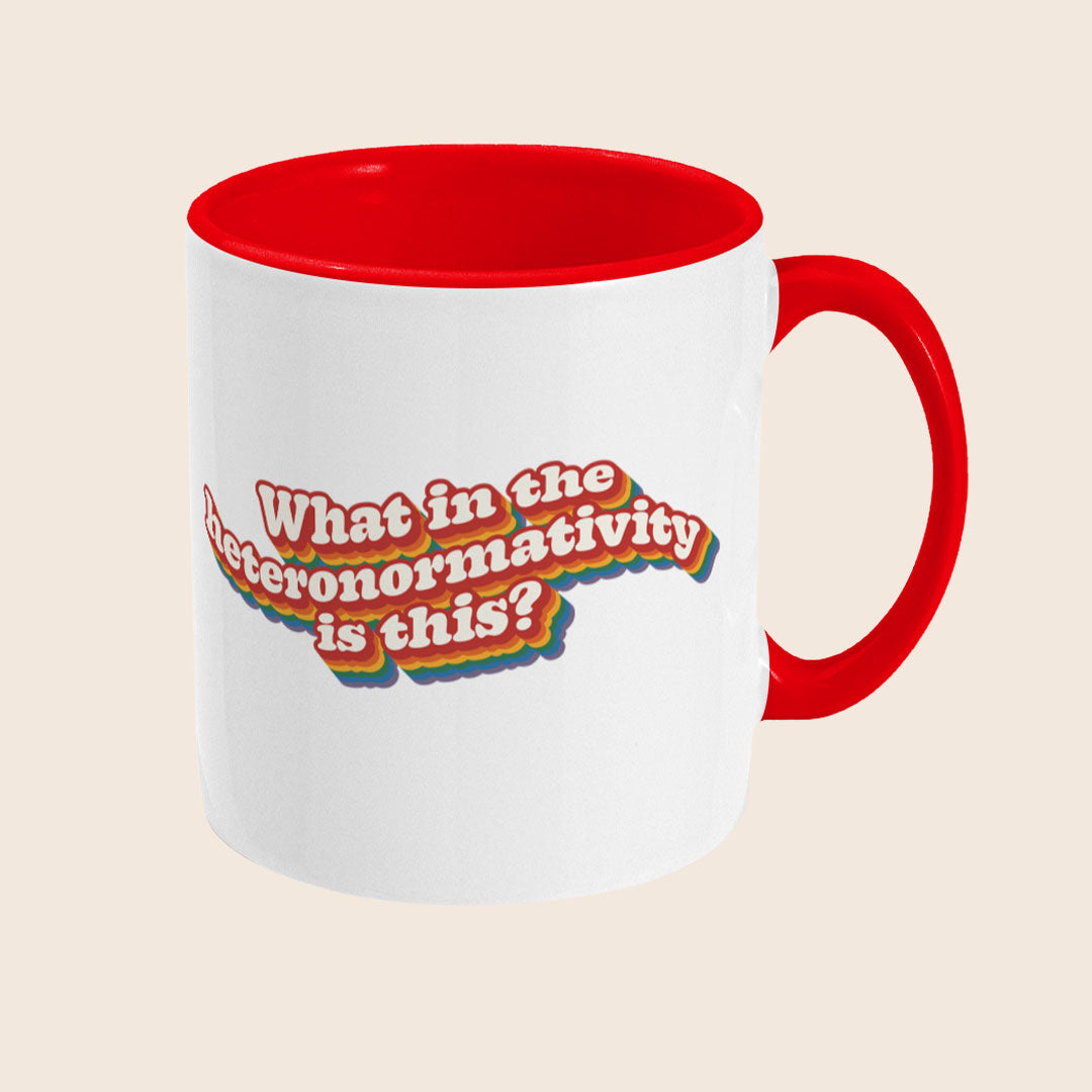 White ceramic mug with a red handle and red interior shown from a right angle, displaying the rainbow retro text reading “What in the heteronormativity is this?”.