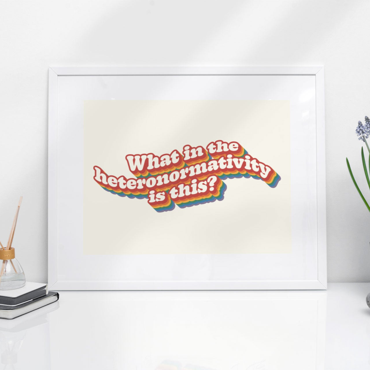 A framed print featuring the phrase What in the heteronormativity is this? with a retro rainbow outline, displayed on a white desk next to a potted plant.