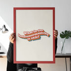 A person holding a large framed What in the heteronormativity is this? print, sitting at a desk, partially obscured behind it.