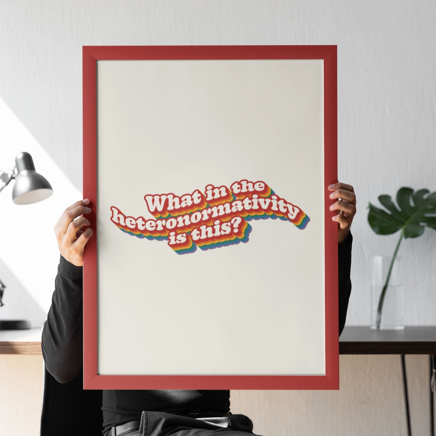 A person holding a large framed What in the heteronormativity is this? print, sitting at a desk, partially obscured behind it.