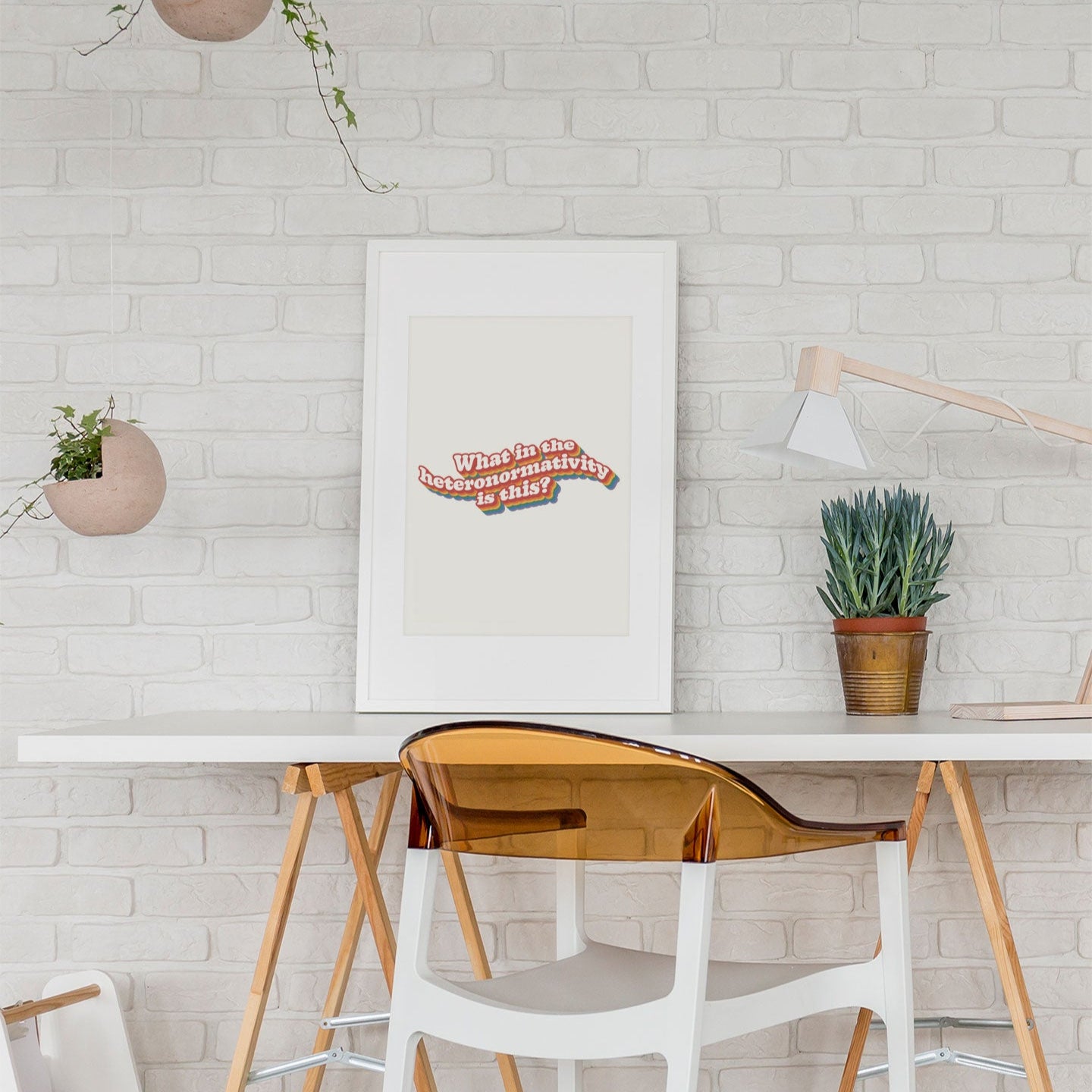 A bright, minimalist workspace with a framed print of What in the heteronormativity is this? displayed on a white brick wall.