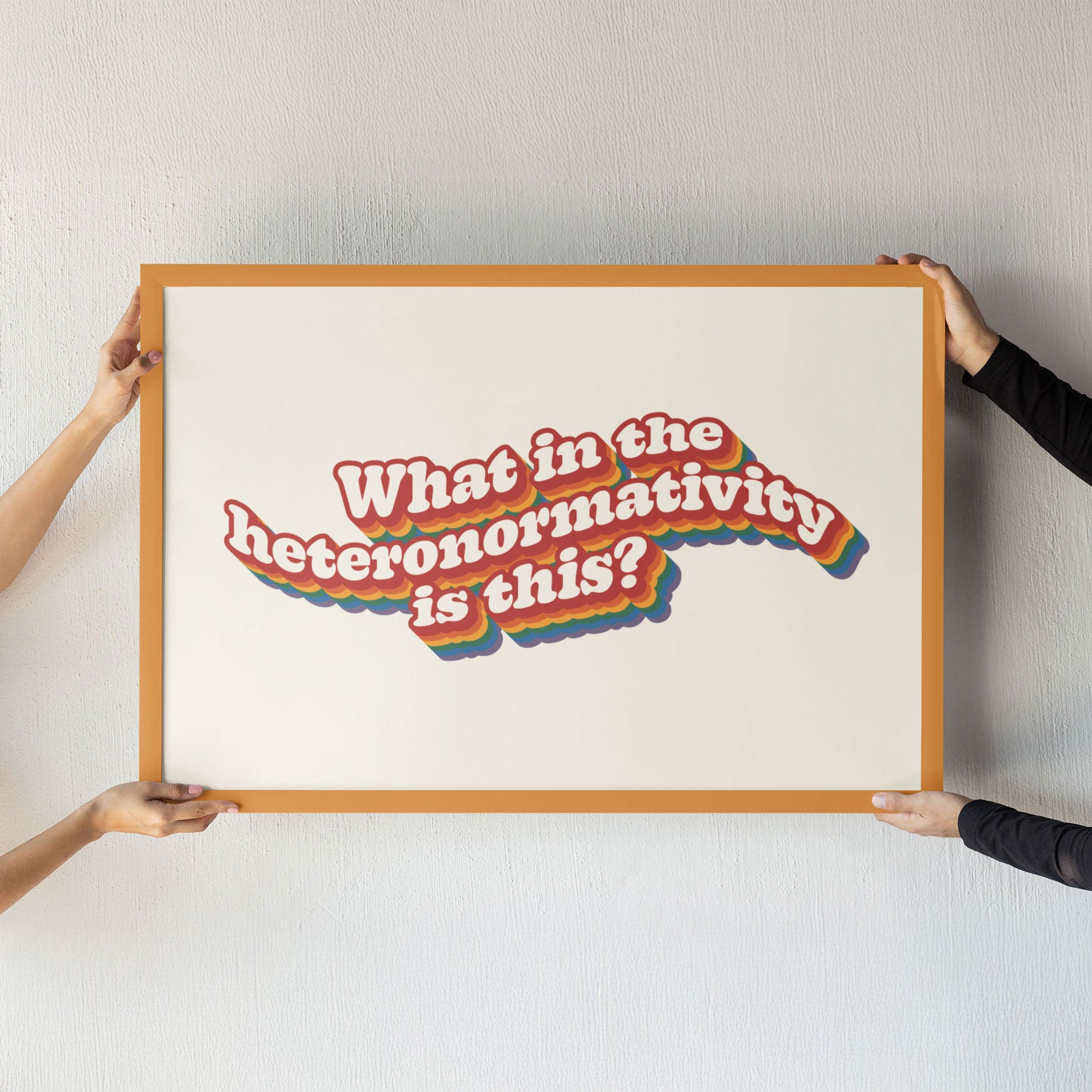 Two people holding up a large framed What in the heteronormativity is this? print, showcasing its bold typography and vibrant rainbow accents.