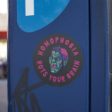 Load image into Gallery viewer, A round black sticker with a vibrant zombie illustration in the centre. The zombie&#39;s face is green and blue with purple hair, an exposed brain, and red eyes. The text around the zombie reads &quot;HOMOPHOBIA ROTS YOUR BRAIN&quot; in pink, bold, dripping letters. The sticker is placed on a blue metal surface.