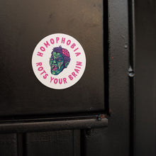 Load image into Gallery viewer, A round white sticker featuring a colourful zombie face in the centre. The zombie is illustrated with a green and blue face, purple hair, an exposed brain, and red eyes. Surrounding the zombie is the text &quot;HOMOPHOBIA ROTS YOUR BRAIN&quot; in pink, bold, dripping letters. The sticker is placed on a black wooden surface.