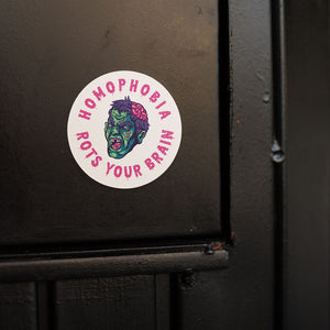 A round white sticker featuring a colourful zombie face in the centre. The zombie is illustrated with a green and blue face, purple hair, an exposed brain, and red eyes. Surrounding the zombie is the text "HOMOPHOBIA ROTS YOUR BRAIN" in pink, bold, dripping letters. The sticker is placed on a black wooden surface.