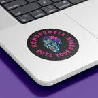 A circular sticker with the text "HOMOPHOBIA ROTS YOUR BRAIN" in bold, pink, jagged letters is placed on the bottom corner of a silver laptop. The sticker features a graphic of a zombie-like face with green skin, red eyes, and an exposed pink brain, designed to convey an anti-homophobia message. The laptop is set against a purple background.