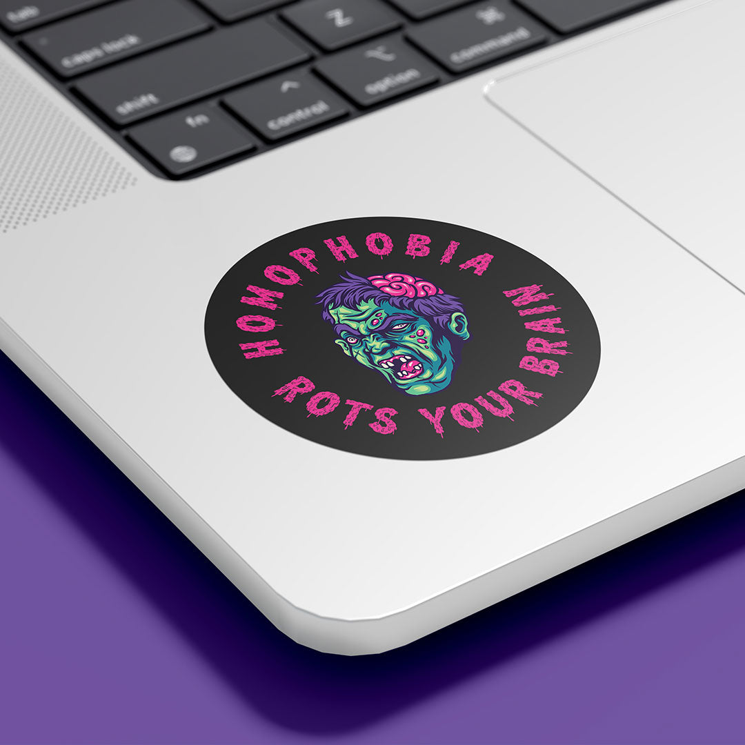 A circular sticker with the text "HOMOPHOBIA ROTS YOUR BRAIN" in bold, pink, jagged letters is placed on the bottom corner of a silver laptop. The sticker features a graphic of a zombie-like face with green skin, red eyes, and an exposed pink brain, designed to convey an anti-homophobia message. The laptop is set against a purple background.
