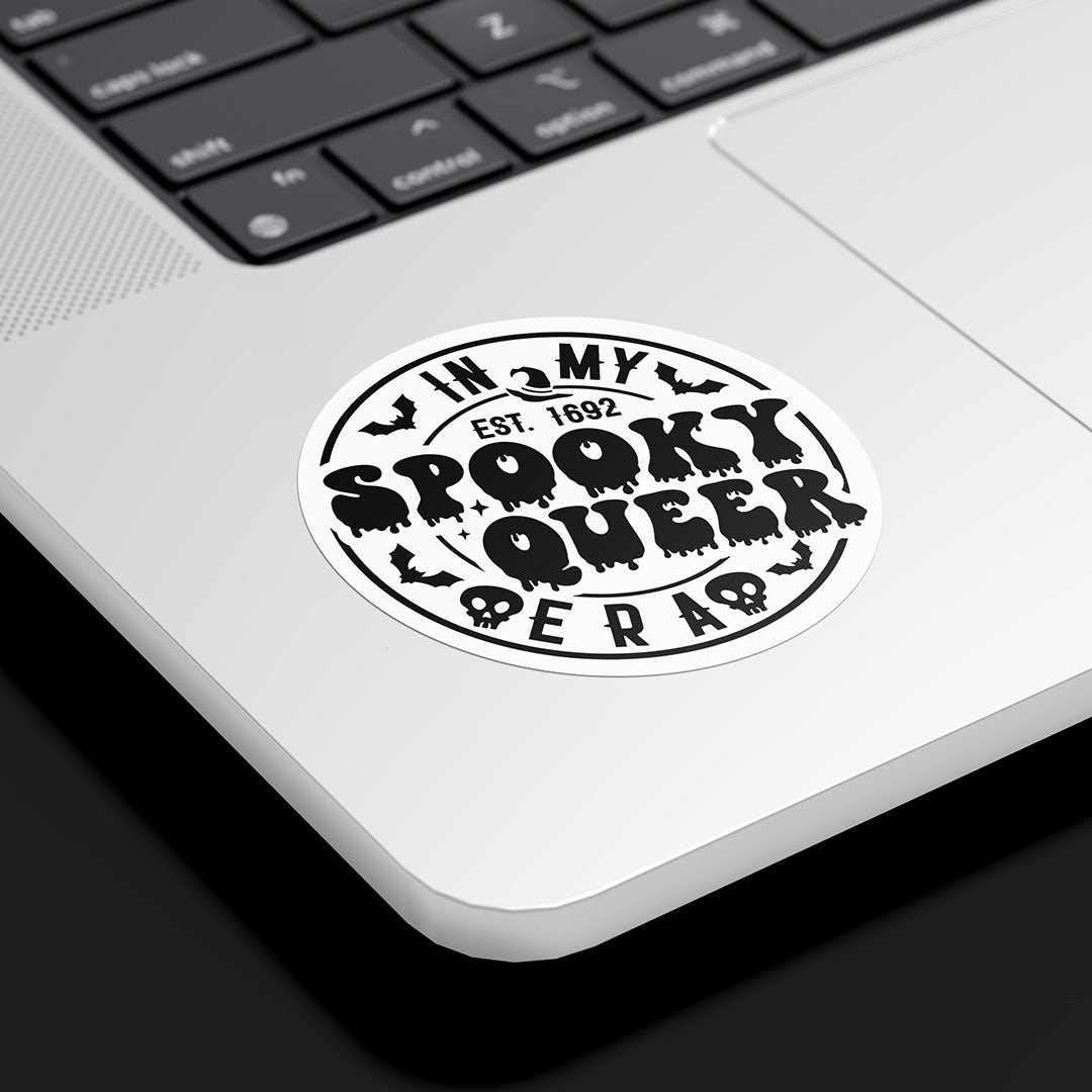 A round sticker with the text "In My Spooky Queer Era, Est. 1692" is affixed to the bottom corner of a silver laptop. The design is in black and white, featuring dripping letters, bat silhouettes, and small skull icons, giving it a spooky, vintage Halloween vibe. The laptop rests on a sleek black surface, highlighting the sticker's bold, monochromatic design.